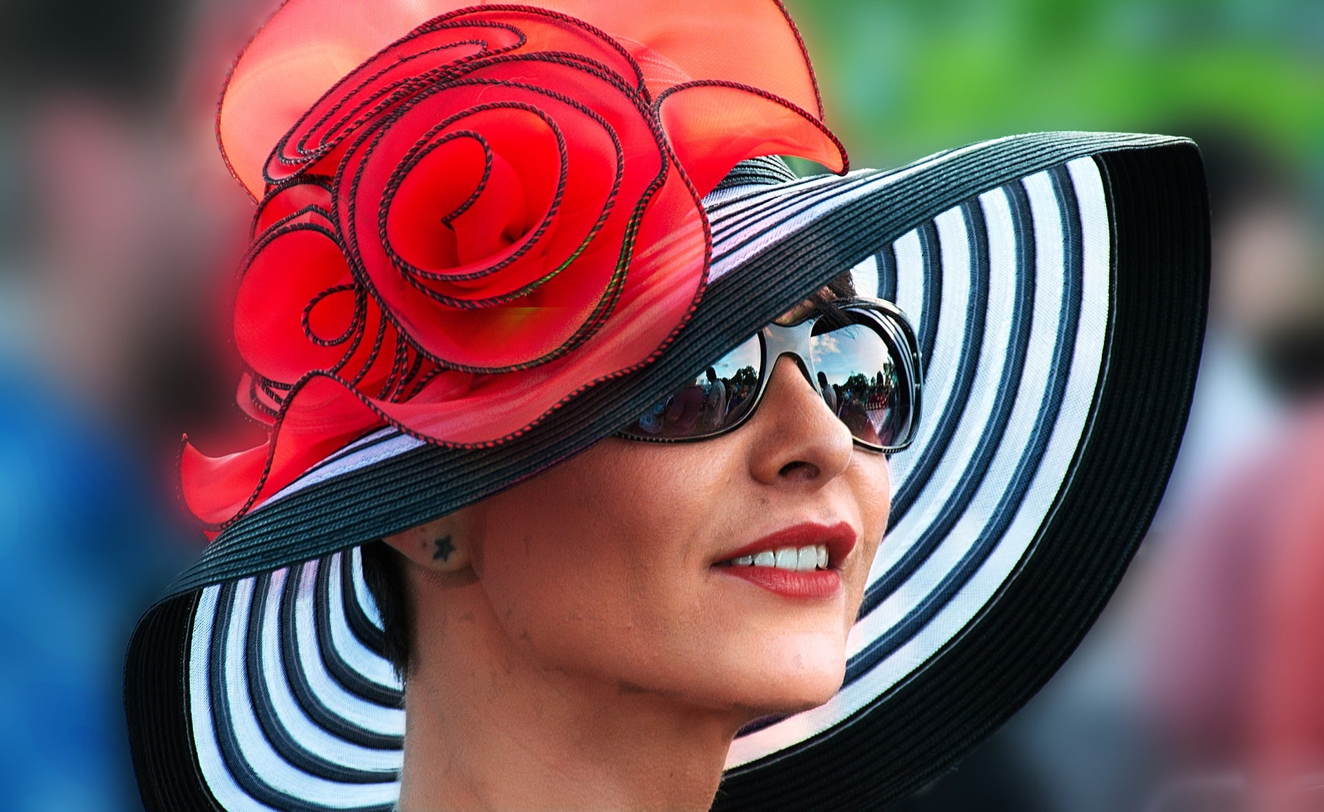 Woman wears a lavish black and white hat with red roses, and sunglasses.
